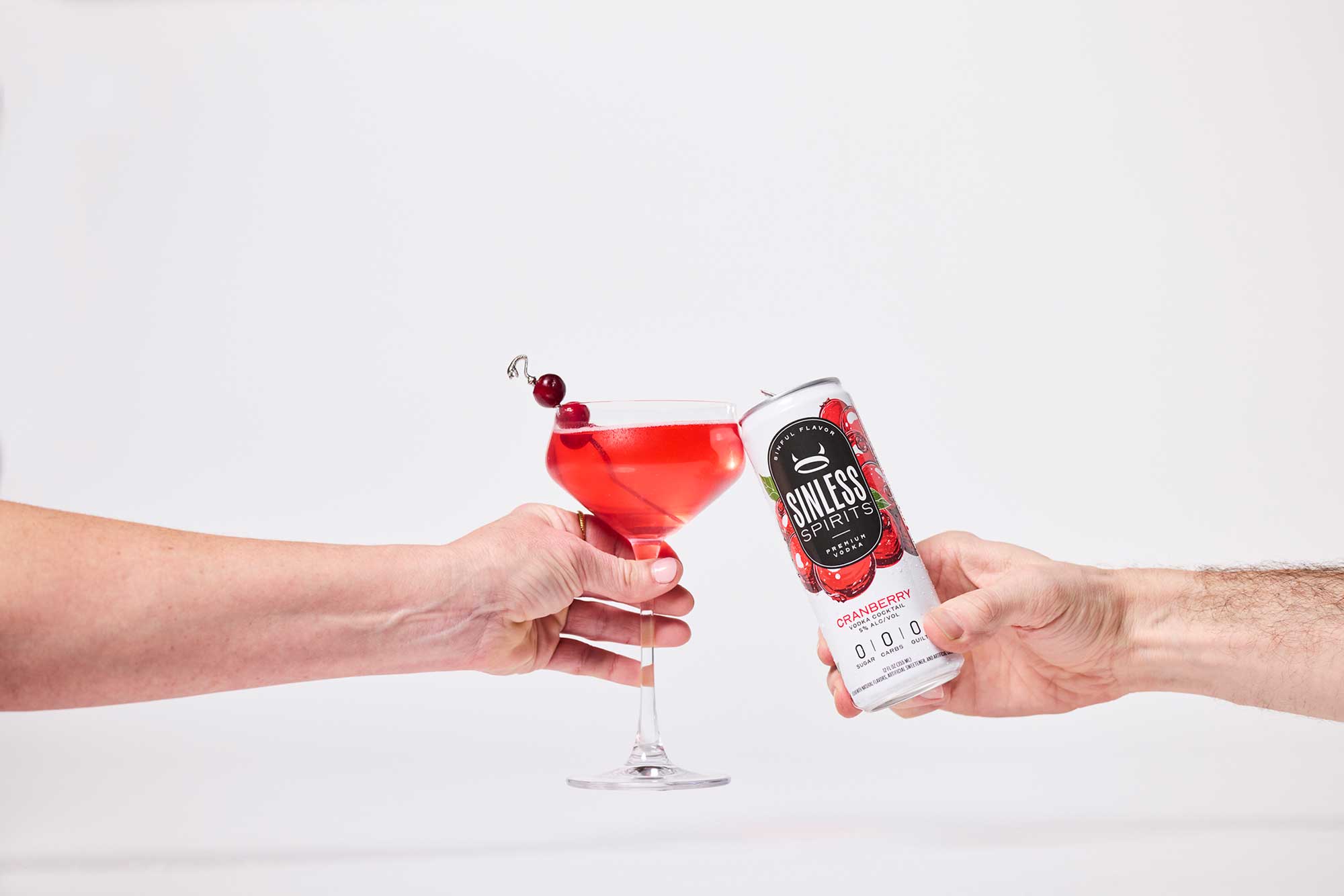 two hands clinking a glass and a can of sinless spirits cranberry