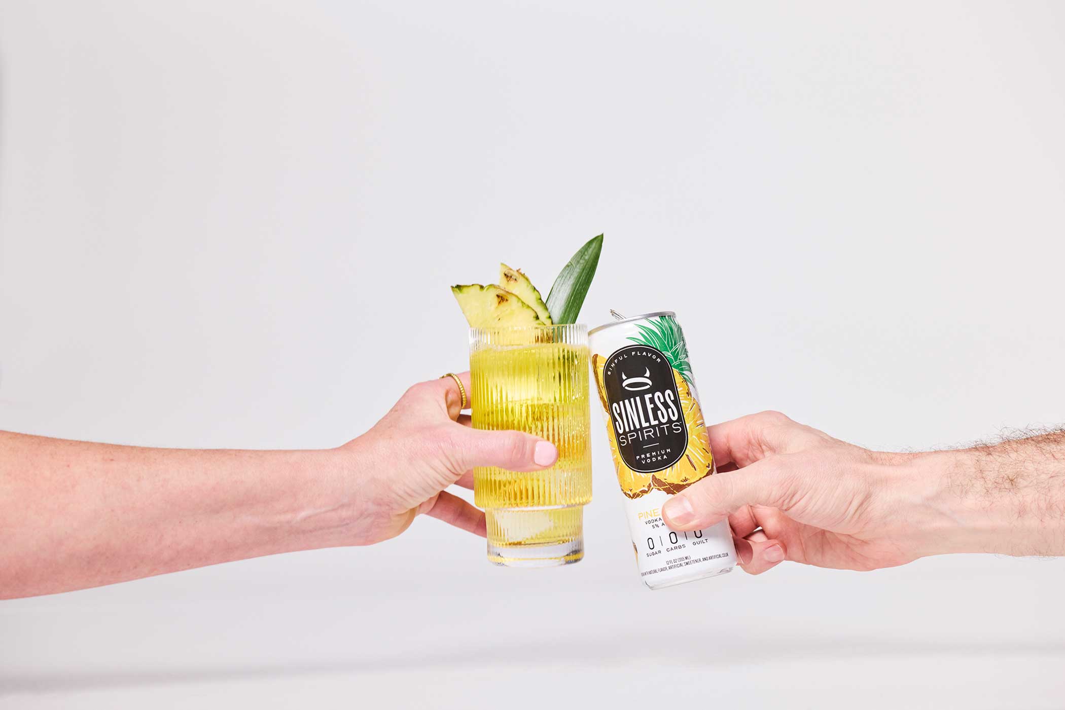 two hands clinking a glass and a can of sinless spirits pineapple