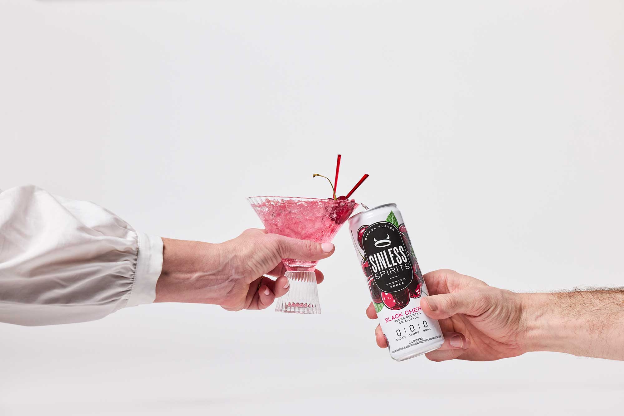 two hands clinking a martini glass and a can of sinless spirits black cherry