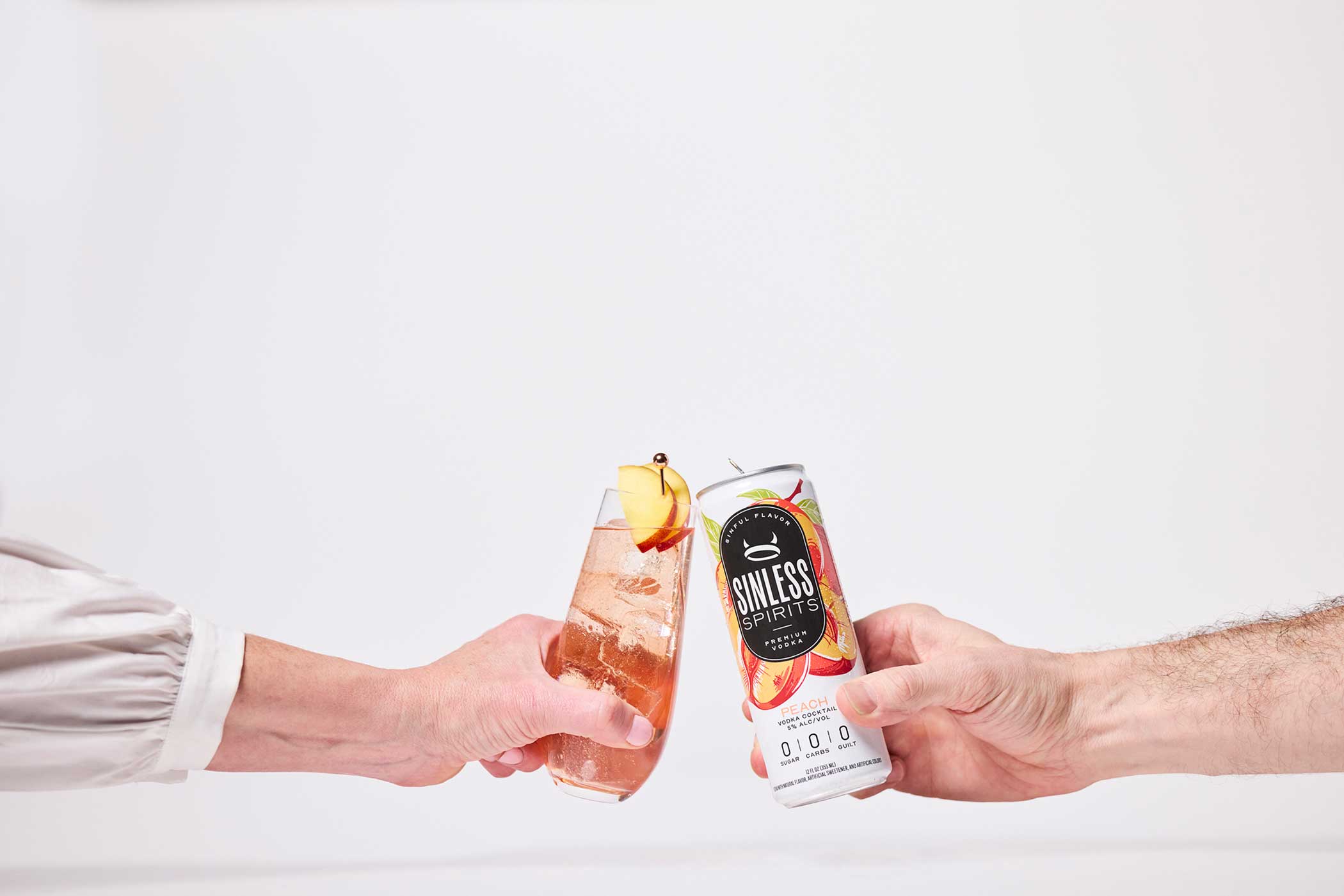 two hands clinking a glass and a can of sinless spirits peach