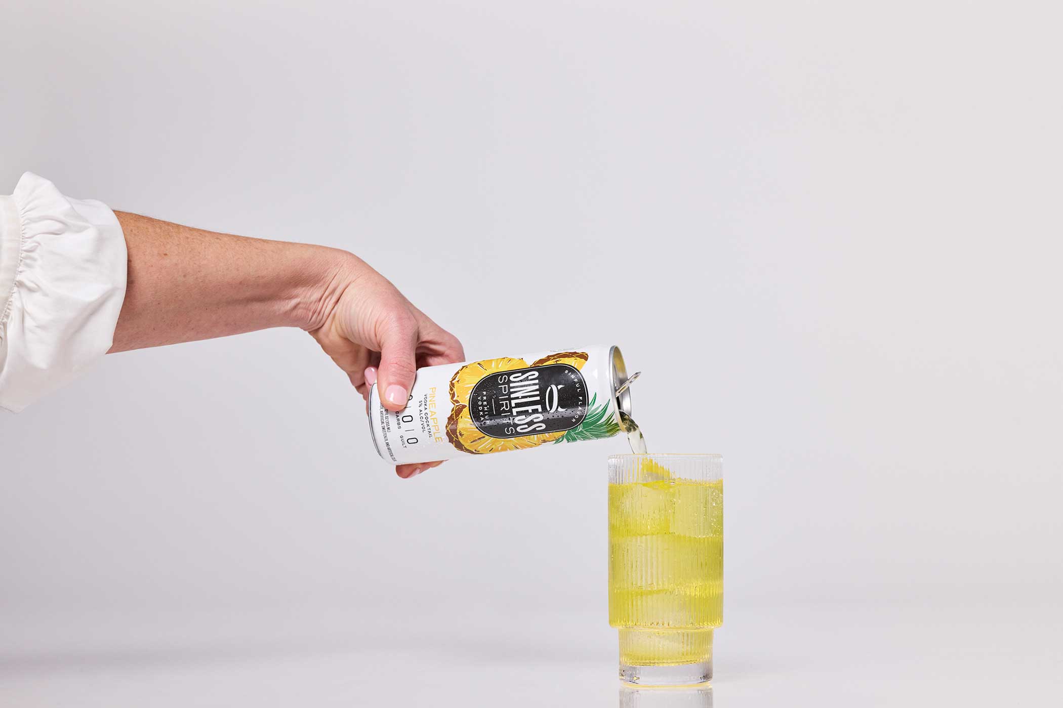 someone pouring a can of sinless spirits pineapple into a ribbed glass with ice